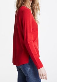 Roter Strickpullover mit figurnaher Passform und oversized Ärmeln, der eine horizontale Naht über der Brust und einen etwas längeren Saum hinten aufweist.