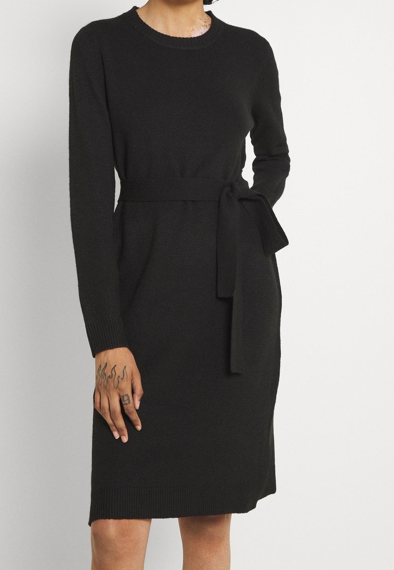 Černé pletené šaty s dlouhými rukávy, kulatým výstřihem a páskem v pase. Mají hřebíkované manžety a spodek, hladkou texturu.