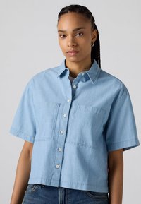 Camicia azzurra a maniche corte in denim, con chiusura a bottoni frontale, due tasche sul petto e orlo corto.