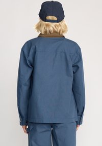 Blaues Denim-Jacken mit lockerem Schnitt, langen Ärmeln, dunklen Knöpfen und abgerundetem Saum. Getragen mit einer passenden blauen Kappe.