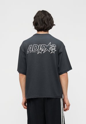 Άτομο με μαύρα μαλλιά που φοράει μια μαύρη υπερμεγέθη μπλούζα Adidas Originals με λευκά γραφικά αστεριών και λογότυπο στην πλάτη.