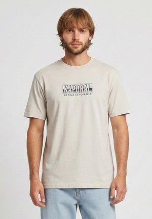 Uomo con capelli castano chiaro e barba che indossa una t-shirt beige "Kaporal" e jeans azzurri, in piedi contro uno sfondo chiaro e uniforme.