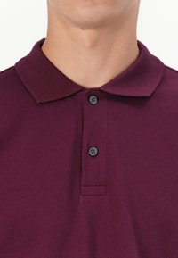 Burgundy polo skjorte lavet af tekstureret stof, med en klassisk krave med to sorte knapper og korte ærmer.