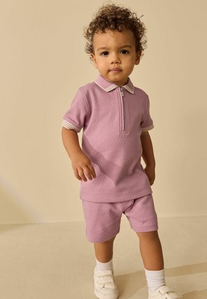 Toddler with curly hair wearing a pink short-sleeve zip-up shirt, matching shorts, white socks, and white shoes against a beige background.