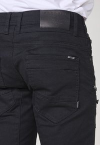 Črne jeans hlače z teksturirano površino, ki vključujejo več žepov, usnjen etiketi na pasu ter kontrastne šive.