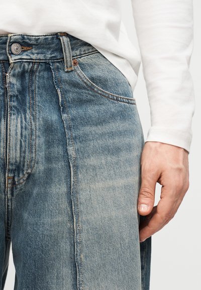 Κοντινό πλάνο ενός ατόμου που φοράει ξεθωριαμένα μπλε τζιν και λευκό πουκάμισο με μακριά μανίκια, με το δεξί του χέρι να ακουμπά απαλά στο πλάι.