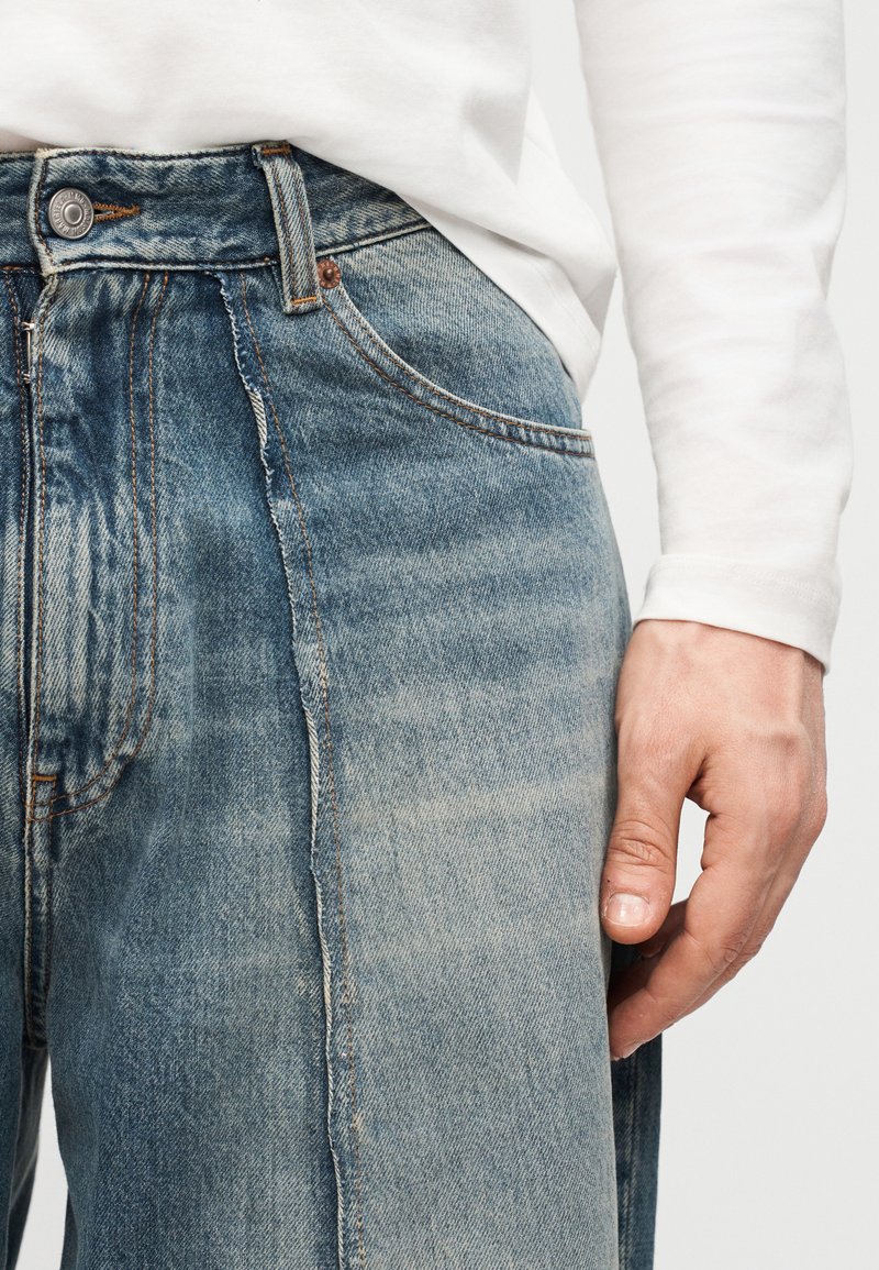 Close-up van een persoon die verwassen blauwe spijkerbroek en een wit shirt met lange mouwen draagt, met de rechterhand licht langs het lichaam rustend.