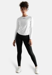 Langærmet hvid atletisk top og sorte leggings, der har en tætsiddende pasform, glat tekstur og logo-detaljer. Fodtøjet er lyse sneakers.