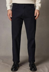 Navy cotton trousers with a straight fit, featuring belt loops, front pockets, and a button closure, paired with black leather shoes.