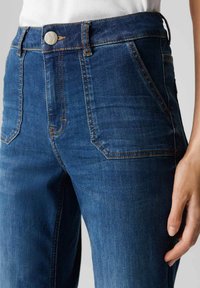 Jeans a vita alta blu scuro realizzati in denim, dotati di tasche frontali, cuciture arancioni e chiusura con bottone in metallo. Texture liscia con leggero elastico.