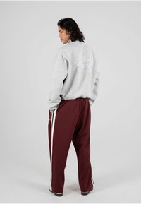 Felpa grigia con dettagli ricamati sulla schiena, abbinata a pantaloni larghi bordeaux con strisce bianche sui lati. Tessuto complessivamente liscio.