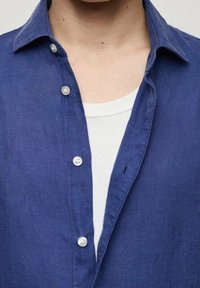 Chemise en lin bleu marine avec col pointu, boutonnage devant avec des boutons blancs, portée sur un t-shirt blanc à col rond.