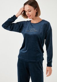 Navy sininen velour-loungewear-setti, jossa on pitkät hihat, kultakirjaillulla tekstillä "sunrise candle co" ja rento istuvuus. Pehmeä rakenne.