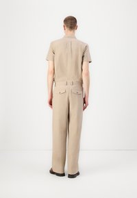 Cămașă bej cu mânecă scurtă și pantaloni largi din țesătură texturată. Pantalonii au două buzunare la spate cu închideri cu buton.
