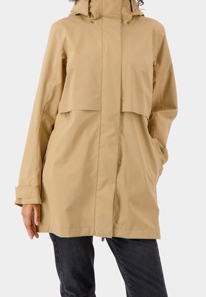 Imperméable beige à capuche avec une fermeture éclair frontale et un rabat, porté avec un pantalon sombre ; main gauche du modèle dans la poche, main droite relâchée le long du corps.