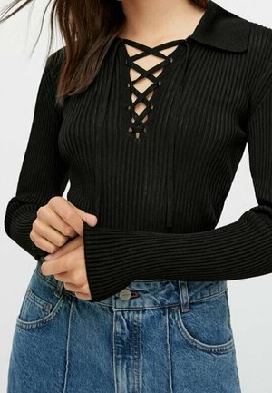 Femme portant un top noir côtelé à manches longues avec un col lacé en croisillons et un jean bleu taille haute.