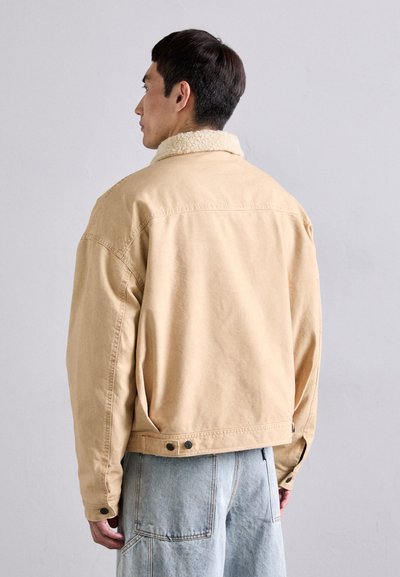 Beige denim jakk, millel on villane krae, suured tagataskud ja nööpdetailid. Üks suurus liiga suur sobivus ning nähtavad õmbluse detailid.