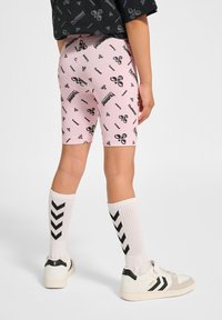 Roze fietsbroekjes met een herhaald patroon van zwarte logo's, witte kniekousen met zwarte chevronstrepen en zwart-witte sneakers.