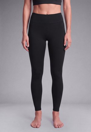 Mujer que lleva leggins negros de cintura alta y un sujetador deportivo negro, de pie descalza frente a un fondo gris liso.