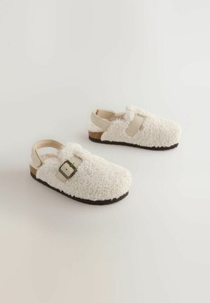 Sabots en shearling blanc avec une surface texturée, dotés d'une boucle latérale et d'une semelle en caoutchouc foncé. Design ouvert à l'arrière pour un enfilage facile.