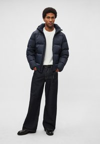 Casaco acolchoado azul-marinho com capuz, com secções acolchoadas, usado por cima de uma camisa branca e jeans escuros de perna larga. Combinado com sapatos pretos.