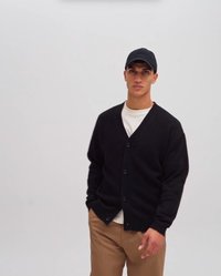 Ung mand iført en sort kasket, sort knappet cardigan, hvid t-shirt og brune bukser, stående mod en ensfarvet lys baggrund.