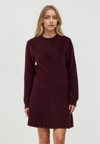 Burgunderfarbener Pullover aus Strick mit ribbeligem Rundhalsausschnitt, langen Ärmeln und einem passenden plissierten Rock, der eine weiche Textur und eine figurbetonte Silhouette zeigt.