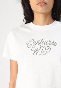 Valkoinen puuvillapaita, jossa on vihreä kirjailtu logomuotoilu, joka lukee "Carhartt WIP". Lyhyet hihat ja klassinen pyöreä kaula-aukko.