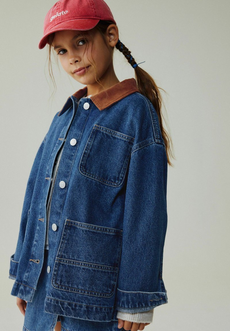 Jachetă din denim cu guler din catifea maro, nasturi argintii, două buzunare frontale și croială lejeră. Însoțită de o șapcă roșie cu text alb.