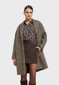 Vrouw in een gebloemde blouse, een bruine corduroy rok, doorschijnende panty’s, kniehoge laarzen en een geweven bruine oversized jas, staand met één hand in haar zak.