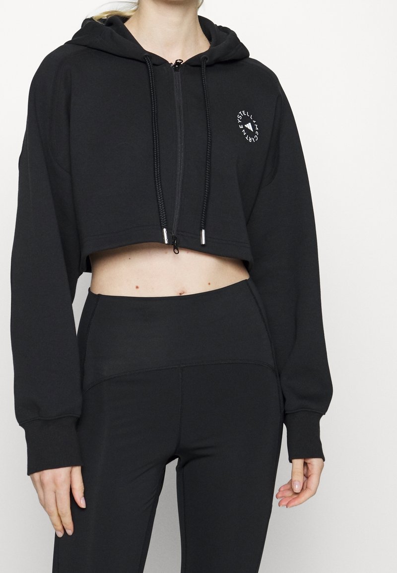 Femme portant un sweat à capuche court noir avec un logo circulaire et un legging noir taille haute, debout devant un fond blanc uni.