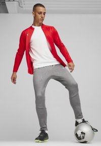 Rode sportjack, witte shirt en grijs gestreepte broek gedragen met zwarte voetbalschoenen. Zwarte en zilveren voetbal op de grond.