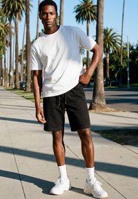 Man draagt wit t-shirt, zwarte korte broek, witte sokken en witte sneakers, staand op een trottoir met palmbomen aan weerszijden bij daglicht.