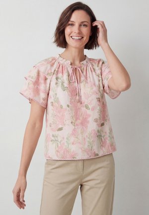 Donna sorridente con indosso una camicetta floreale rosa chiaro con maniche corte arricciate e pantaloni beige su uno sfondo chiaro uniforme.