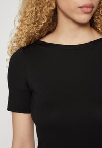 Černý top s krátkým rukávem a širokým dekoltem, vyrobený z hladkého, elastického materiálu. Jednoduchý design a přiléhavý střih, který ukazuje jemnou texturu.