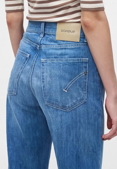 Personne portant un jean en denim bleu avec une étiquette en cuir "DONDUP" sur la ceinture et une chemise beige à rayures.