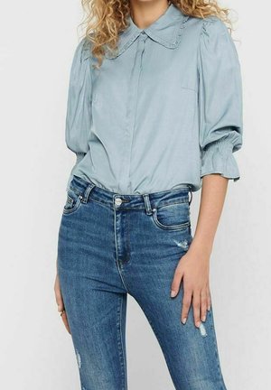Vrouw met krullend haar draagt een lichtblauwe blouse met pofmouwen, ingestopt in aangesloten blauwe jeans met lichte slijtage, staand tegen een effen achtergrond.