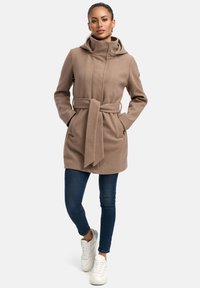 Beige wool blend coat with a high collar, belt, and side pockets; worn over dark jeans and white sneakers. Smooth texture and tailored fit.