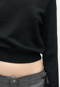 Κοντινό πλάνο ενός ατόμου που φοράει μαύρο κοντό πουλόβερ και γκρι παντελόνι, με ένα μικρό εκτεθειμένο τμήμα της μέσης.