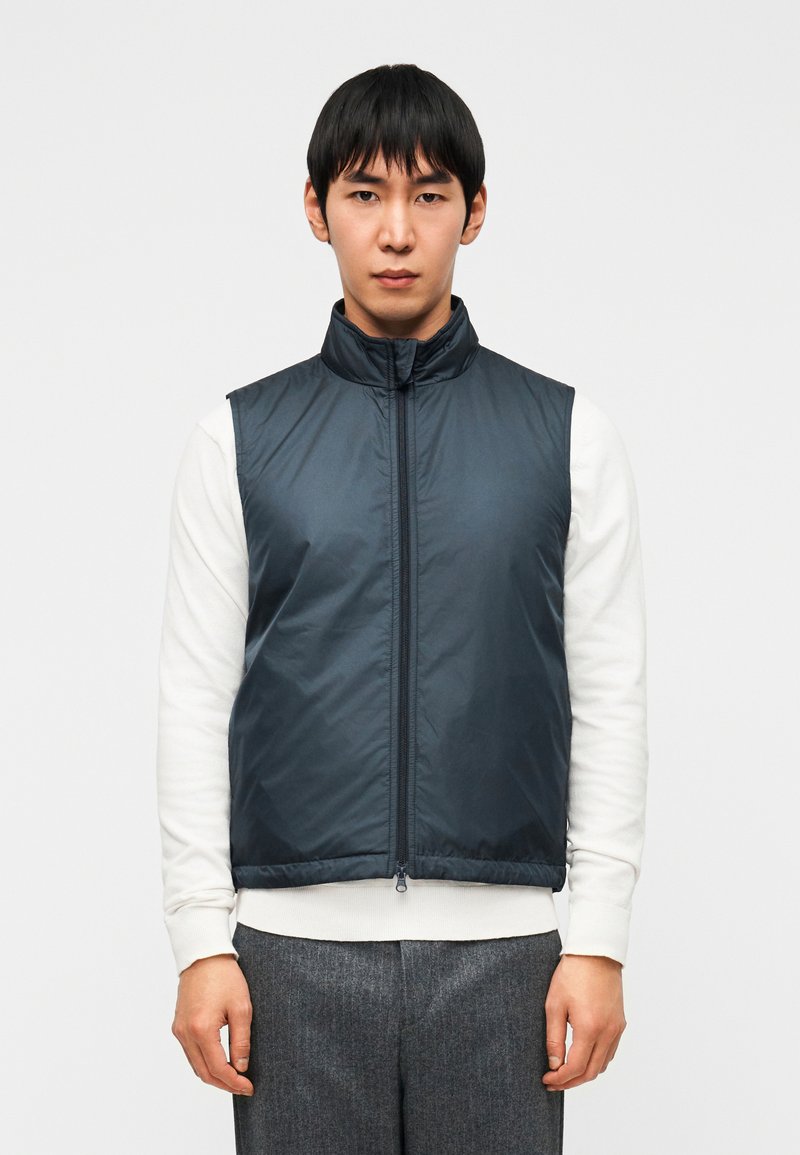 Σκούρο μπλε μπουφάν αμάνικο με υψη λό γιακά, μπροστινό φερμουάρ και σχεδιασμό χωρίς μανίκια, φορεμένο πάνω από λευκό πουκάμισο με μακριά μανίκια. Ομαλή υφή υφάσματος.