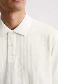 Witte polo-shirt met wafelstructuur, voorzien van een kraag, een knoopsluiting met twee knopen, en zachte stof. Het ontwerp is eenvoudig en klassiek.