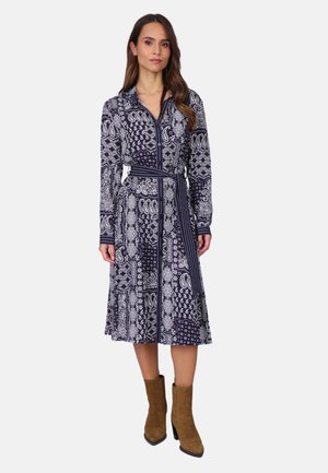 Femme portant une robe midi à manches longues, à motifs bleu marine et blanc, avec une ceinture à nouer à la taille, associée à des bottines marron en daim à talons montant jusqu'au milieu du mollet.