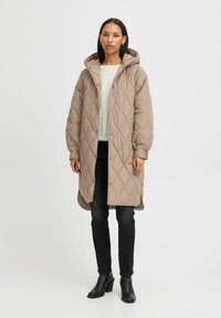 Manteau long matelassé beige avec capuche, doté de fermetures à pression, de poches latérales et d'une texture douce, associé à un pull léger et un pantalon noir.