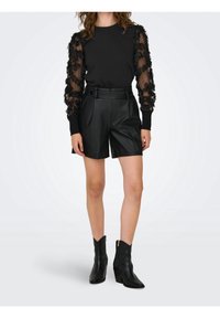 Suéter negro con mangas transparentes y con volantes, combinado con shorts negros de cuero sintético de talle alto y botines negros de tacón.