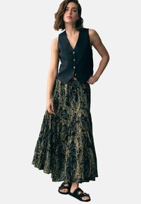 Schwarze Leinenweste mit goldenen Knöpfen, kombiniert mit einem fließenden Maxirock in Schwarz und Goldmuster und schwarzen Sandalen mit Nieten.