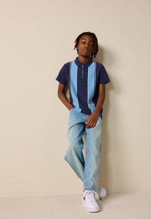 Garçon avec des dreadlocks courtes appuyé contre un mur beige, portant un polo rayé bleu marine et bleu clair, un jean bleu clair et des baskets blanches.