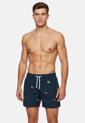 Uomo muscoloso che indossa pantaloncini da bagno blu navy con piccole stampe a fette di anguria e un laccio bianco su uno sfondo semplice.