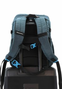 Mochila en azul verdoso con correas acolchadas y acentos negros, unida a una maleta negra. Las características incluyen hebillas de plástico azul y un respaldo de malla.
