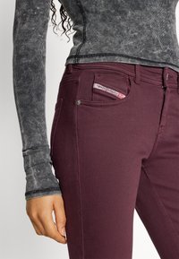 Vaqueros burdeos de corte slim, con un pequeño logo en el bolsillo delantero y detalles sutiles de costura. Camiseta de manga larga gris con textura acanalada.