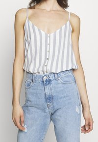 Randig ärmlös topp med v-ringning; ljusgrå och vita färger; har små knappar fram och en avslappnad passform. Bärs med blå jeans.